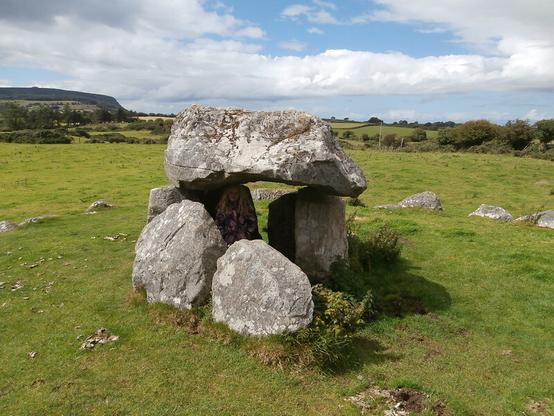 A small dolmen-style burial chamber with a chunky capstone and four orthostats. A small child (she was nine, I think) stands inside the chamber. You can see the circle of kerb of low stones that surrounds the burial chamber. Blue sky, white clouds, green grass.