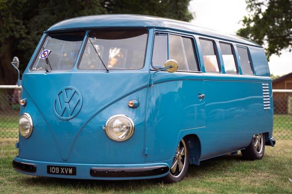 A beautiful blue split screen Volkswagen Camper Van, surrounded by a lush green lawn. The classic van boasts a solid blue exterior and a split screen design. It has round headlights, a sloping hood, and a curved windscreen that are signature features of the model. The recognizable VW emblem is prominently displayed on the front grille, and the rear opens up to reveal a compact and functional living space.