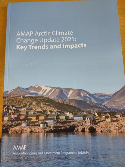 Photo of a report called AMAP Arctic Climate Change Update 2021 Key trends and impacts a picture of bare mountains in a blue sky with a small colourful town on the rocky shore below. The sea in front is calm.