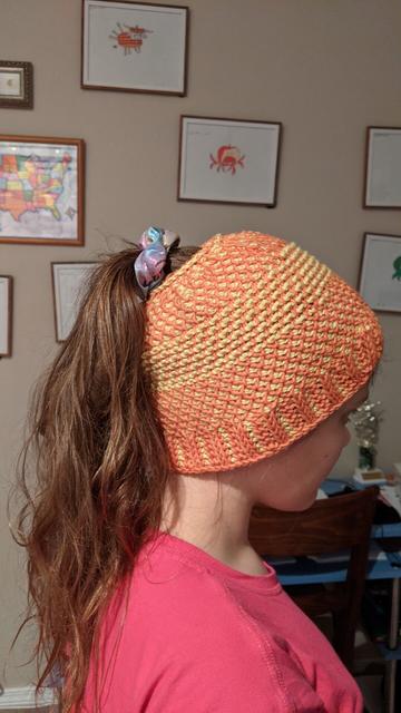 Side view of yellow and orange pony tail hat with layers of different stitches - some like lattice, some like bumps, some like rubbing, and some hard to describe.