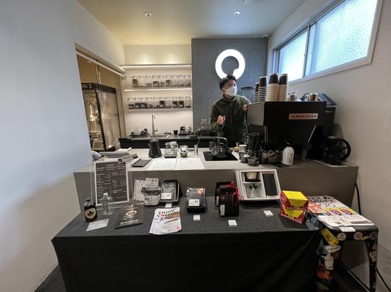 A very crowded coffee counter with espresso machine, jars of beans on the walls, packaged products on a display table. In the far corner in a nook is a roaster. A man prepares coffee behind the counter