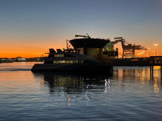 Sunset and light reflecting off the water at the Oakland Jack London Square ferry terminal, with an arriving ferry, the USNS John Glenn towering over it, and shipping cranes in the distance.