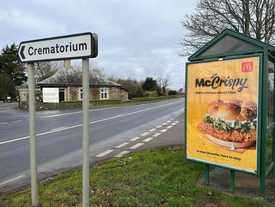 A photo of a road passing by a building in the British countryside. A road sign points toward a crematorium across the street. Slightly behind the sign is a bus stop shelter featuring a McDonalds advertisement for the McCrispy sandwich.