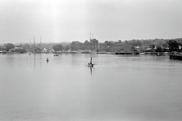 A black and white photograph of a view upriver. Buildings cluster along the shore, and the masts of a large sailing vessel rise behind some buildings. Smaller boats are visible, and a steam-powered launch is heading upriver.