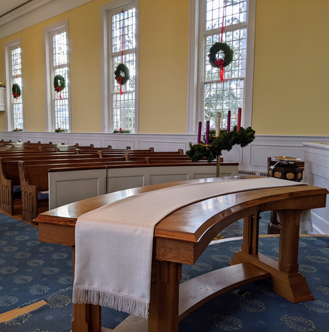 Empty church. A curved, wooden table in the foreground, a wall with 4 tall windows in the background