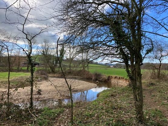 Photo of a river meandering through green fields. The river has cut into the bank on outside of the bend (on the right) leaving a sheer, sandy coloured cliff, which the footpath clings on to! On the inside of the bend is a shallow shingle beach. A few spindly trees line the banks. White clouds in the sky are breaking up revealing large patches of blue, which are reflected in the curve of the river.