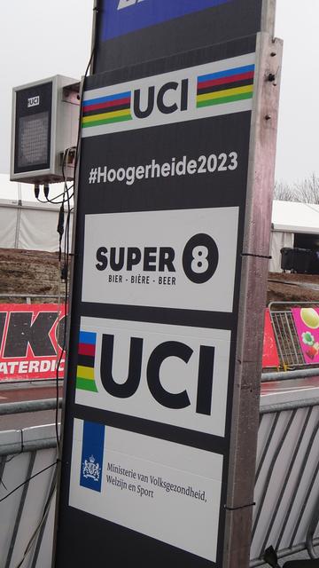 A vertical banner next to the finishing straight at the Hoogerheide Cyclo-Cross World Championships. From top to bottom it has the logos for Santini (clothing), the UCI, #Hoogerheide2023, Super 8 (beer) and the UCI again.