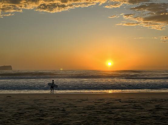 Looking across the beach to see the sun just a bit above the ocean. A lone surfer standing with his board under his arm watch the sunrise. Orange clouds across the top. A small island showing on the left edge.