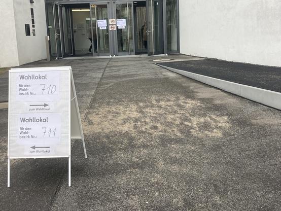 A polling station at a school in Germany. We see the front entrance in the distance. More proximally, a small poster stand informing voters where to go. No people standing in line.