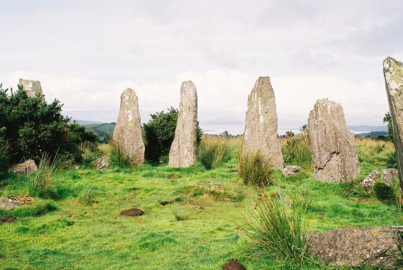 The arc of a stone circle. Six upright, pointed stones (well one of them's a bit stumpier) in a green space, with gorse and reeds and the distant hint of ocean. The sky is grey but the image is quite bright.