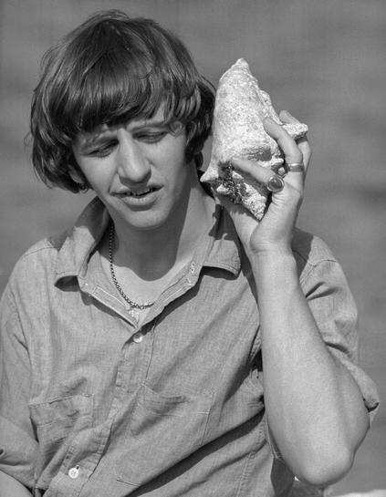 Black and white photo of a mop-haired Ringo Starr in rolled up long-sleeved button-up linen shirt and necklace smiling as he holds a conch shell up to his left ear with his left hand; two large, gaudy rings are visible on his left pinky and left ring finger.