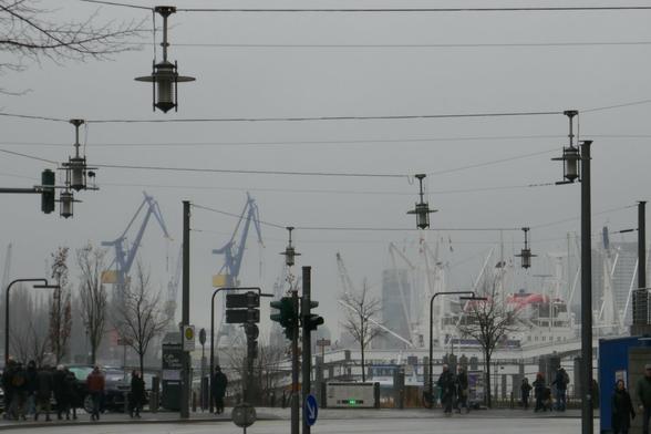 Straßenszene mit Hafenkränen, Masten und Aufbauten von Schiffen im Hintergrund, davor an Seilen zwischen Laternenmasten aufgehängte Straßenlaternen, Passanten auf den Gehwegen am Straßenrand. Bedeckter grauer Himmel.