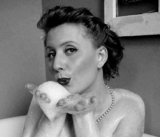 Woman in bathtub with pinup updo, pearl necklace, with hands full of bubble bath suds pretending to blow them at camera.