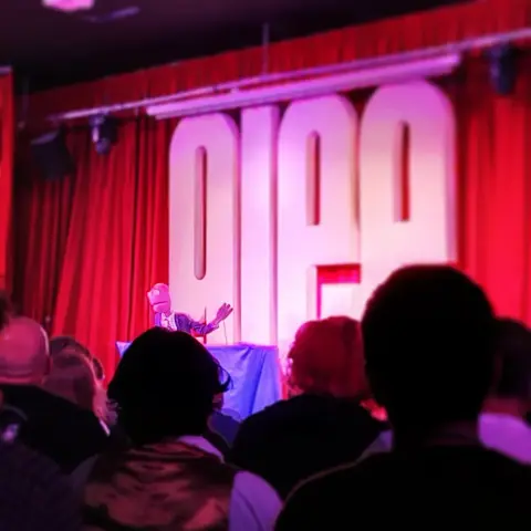 Randy Feltface, a purple puppet in a suit, on stage at a comedy club. In the foreground is an audience and behind Randy is a red curtain and the word GLEE in giant letters.