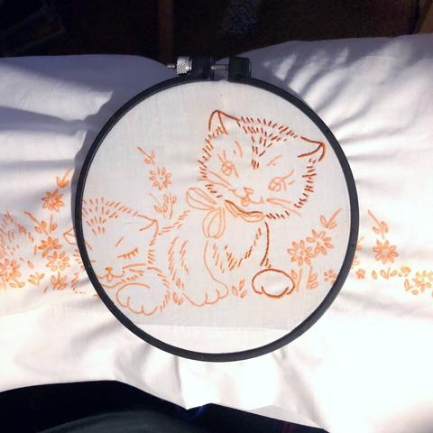 A white pillowcase with a vintage pattern of cats and flowers in a small black hoop. Part of one cat has been done in orange & brown thread