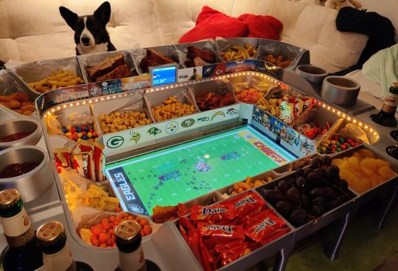 Food-Stadium mit diversen Snacks und zwei Bildschirmen.
Der größere Bildschirm zeigt am Boden des Stadions das NFL-Super-Bowl-Feld mit allen Spielern und Bällen, das kleinere ist in Rang und zeigt vor Spielbeginn die aktuelle Zeit (22:36 Uhr) an.