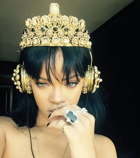 Rihanna's selfie from 2016. She wears a crown & matching headphones. Her hand, with rings on every finger, touches her mouth.