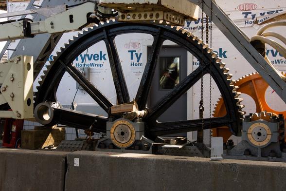 A photograph of a large geared bull wheel of a drawbridge during rehabilitation. Construction materials are around, smaller gears are mounted to the right side of the frame. The bridge operators house is visible in the background, being rebuilt and wrapped in white Tyvek.