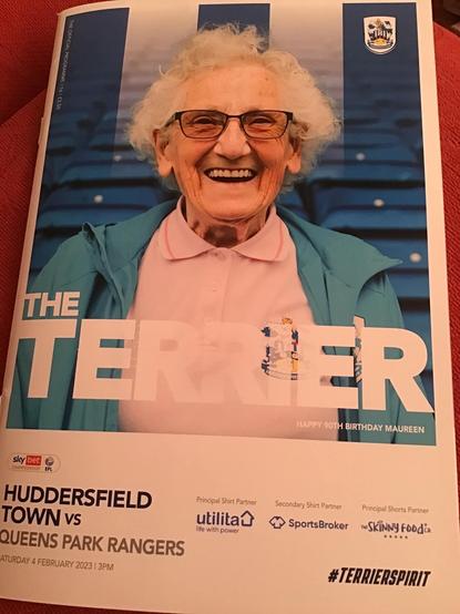 Front cover of the 4 February 2023 match programme for Huddersfield Town Football Club versus QPR. The programme, called The Terrier, features a big picture on most of the front cover of my 90 year old aunt smiling broadly, with the note Happy 90th Birthday Maureen.