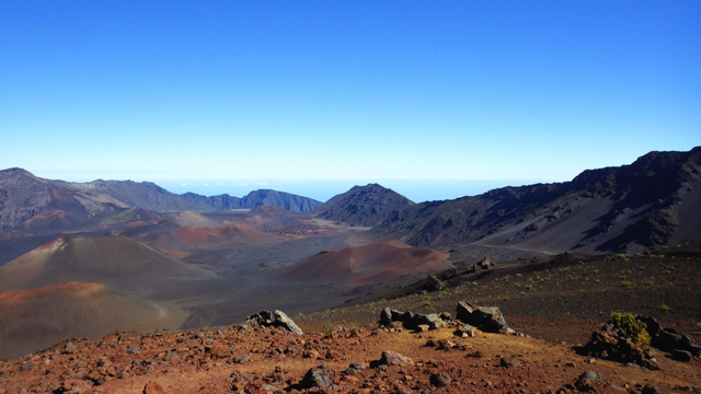 Wide shot of the volcanic crater at Haleakalā there is red soil littered with jagged black rocks in the foreground. there are various cones in the crater -- some are reddish and some are more grey the far side of the crater looks like black/grey mountain peaks.  the background is a very atmospheric blue sky.