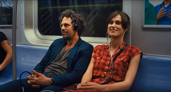 An attractive young white couple in casual 2010s clothing sit on a blue subway seat next to each other, each wearing a pair of headphones (she sports Apple earbuds, he's got over-the-ear cans) plugged into a gargantuan headphone splitter which is in turn plugged into a white Apple iPod. They both are smiling, but she's smiling more.