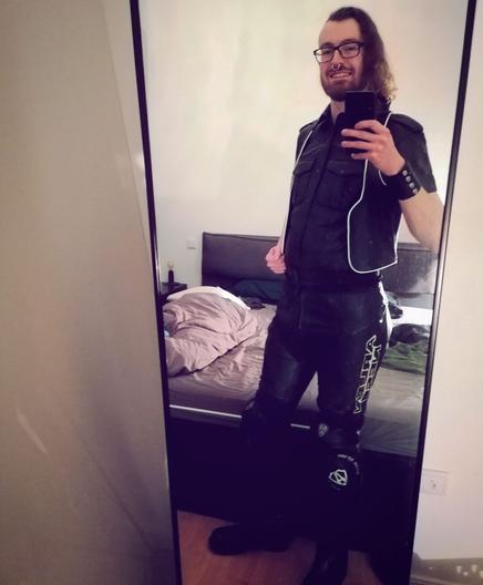 A young man in front of a mirror, dressed in black leather shirt, black and white leather bar vest, a black leather biker pant and leather wristcuff taking a selfie.
