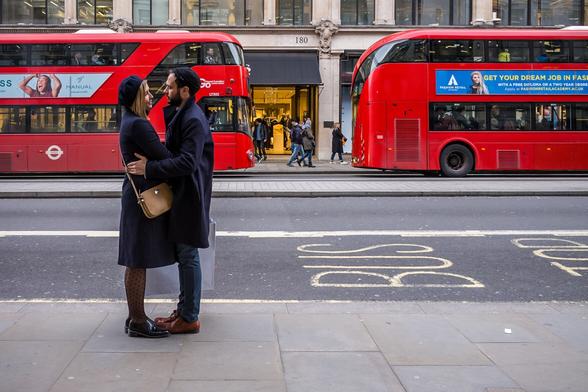 Photograph of a smartly dressed man and woman standing facing each other on a street. In the background are two red London busses. On the road is written the word Bus. They are alone, in the busiest city in the country.