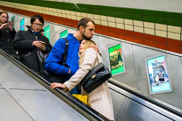 Photograph of a couple riding down an escalator on the London underground. They are on different steps, but holding each other in an embrace. The man rests his chin on the top of her head. He is pouting.