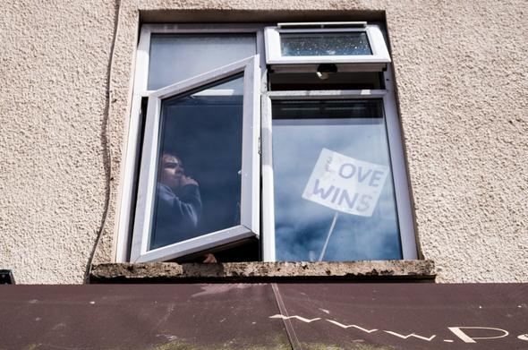 The camera looks up towards a first floor window. Photographed is the window surrounded by a painted pebbledash wall. A person sits looking out of the open half of the window. In the other frame is a small sign reading Love Wins.
