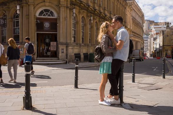 A man and woman kiss on a sunny street bathed in golden light. It looks like a summers evening, he in a short sleeved shirt, she in a skirt.