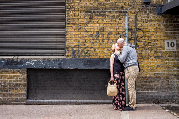 An a late middle aged man and woman kiss on a back street in London. He is holding her neck. She is holding her handbag.