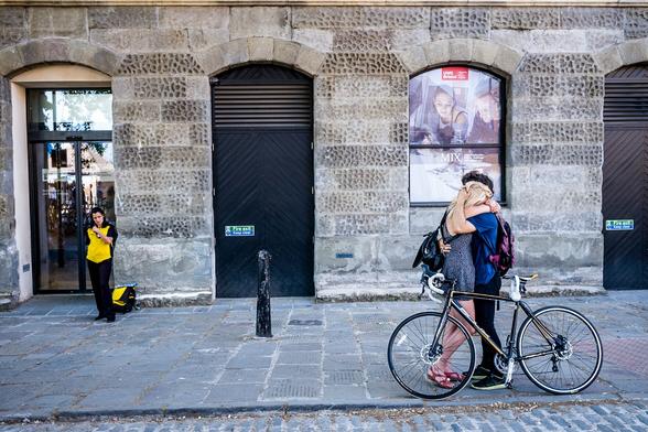 Two people are wrapped in an embrace on the street. They are holding on to each other like their lives depended on it. A bicycle leans against them. In the background a security guard looks on.