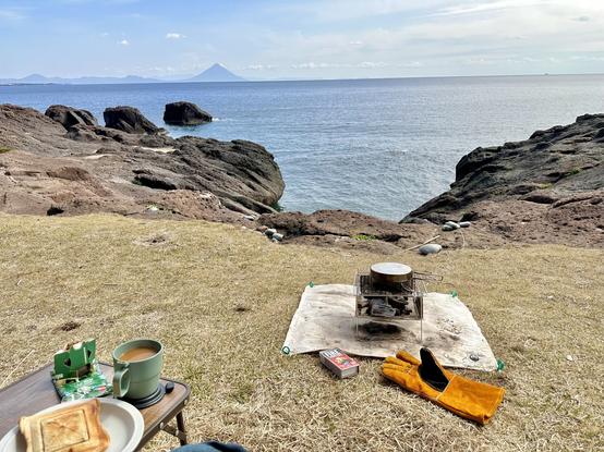 A view of the sea behind some rocks and yellowed grass, with a campfire, coffee and toasted sandwich in the foreground