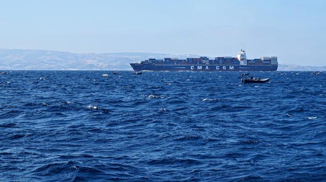 Vom Schiff aus fotografiert. Bewegtes, tief blaues Meer, in der Nähe einige kleine Fischerboote, und weiter hinten ein großes Schiff, voll beladen mit Containern. Ganz im Hintergrund und blass zu sehen die (spanische) Küste.