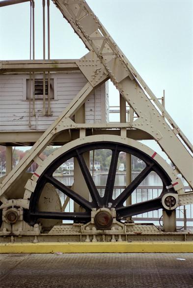 A photo of a large, black, geared bull wheel on a drawbridge. The steel, painted a dingy yellowish color,  shows rust and flaking paint.