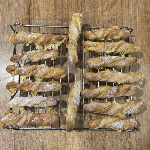 Closeup of fifteen cheese twisters: just before the sticks go into the oven you twist them into a corkscrewish shape, which also encourages some of the  cheese folded into the dough to leak out while they're baking. These still oven hot sticks have a nice gold colour, and the leaked bits of cheese look festive, some yellow, some almost orange (and because I mixed a bit of tandoori powder with the flour I used to cover the dough before cutting it into slices, the sticks have a bit of that red glow too, here and there.