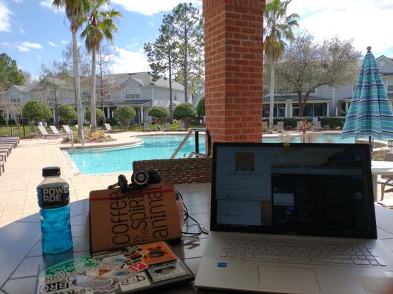 Today's work/lounge area. On a cabana bench centered are two laptops one closed (right) one open. A mountain blast power drink to the left. Behind the closed laptop is a journal that has printed on it "coffee is my spirit animal" Some KOSS Porta Pro headphones above the journal. In the background some palm trees behind the trees is w bright blue sky with puffy white clouds.below is a pool with blue shimmering water. sorry I suck at descriptions.