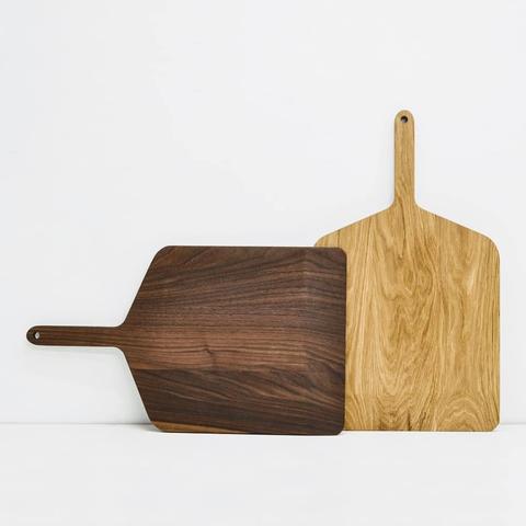 Two pizza paddles. The left is walnut and is laying horizontally. The right is oak and is sitting vertically.