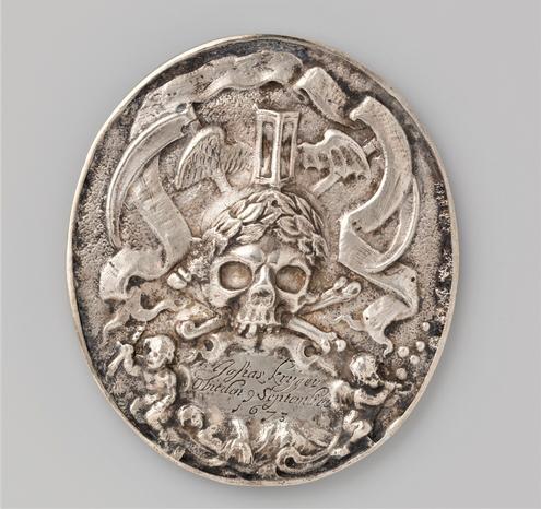 An elliptical silver medal decorated with a skull and crossed bones. Behind the skull is a busy scene featuring reminders of death and the shortness of life such as a winged hourglass, bubbles and scythes.