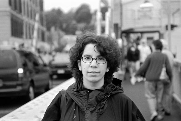 A black and white portrait photograph  (head and shoulders) of a young woman with glasses and medium length dark hair wearing a black coat. She is standing on the sidewalk of a drawbridge, and cars, other pedestrians, and vehicles are visible. The depth of file is shallow, and the background elements are blurred.