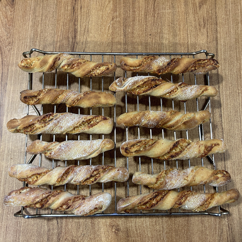 A closeup of 12 twisted cheese sticks, on a cooling rack. Aa nice mix of colours, from the white of the dusted flour, and tge red of tandoori powder, with yellow bits of cheese and the dark honey coloured dough.