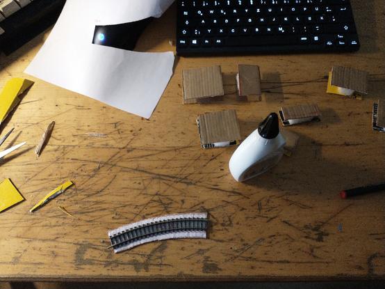 Photo shows the following:

top-view of my workbench, a compact, black pc keyboard without numpad on the top border of the frame, below it there are cardbord squares of various sizes glued together utilizing a small piece of styrofoam inbetween. These are going to be the pillars of an improvised model railroad ramp.