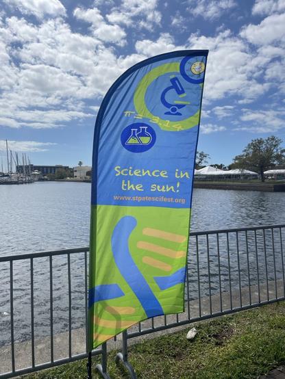 Blue and green flag for the Saint Petersburg Science Festival (https://stpetescifest.org/) that says Science in the sun and shows DNA, flasks, and a microscope. Flag is hung next to the water and the sky behind it is bright blue with white fluffy clouds