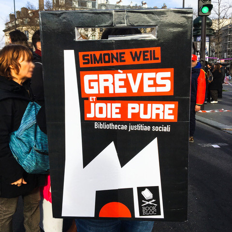 photo de manif, une pancarte reproduisant la couverture de « grèves et joie pure » de Simone Weil : usine blanche sur fond noir, soleil levant rouge dans la porte ouverte.
