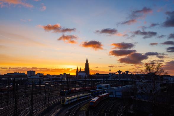 Das Ulmer Münster im Gegenlicht an einem goldenen Morgen mit Wolken. Im Vordergrund der Bahnhof.
