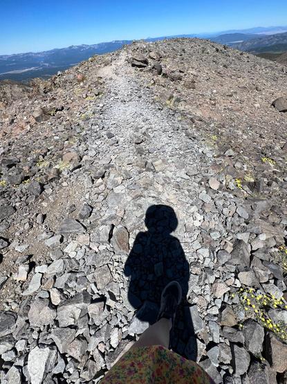 A shadow of me on top of a rocky mountain summit. The sky is blue and there's other mountains on the horizon.