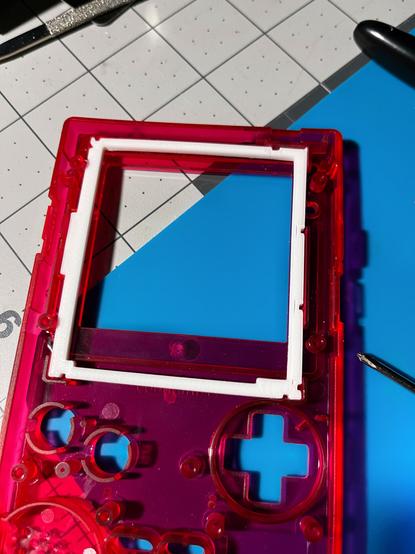 The inside of a transparent Rose Red Game Boy Pocket with a white 3D printed screen brace. It is sitting on a blue silicone mat surrounded by some tools