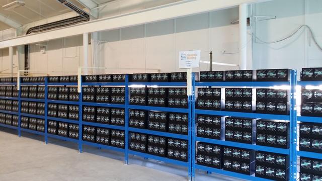 A photo of a Bitcoin mining operation showing racks of computer servers in a warehouse. A sign in front of the servers says "Genesis Mining".