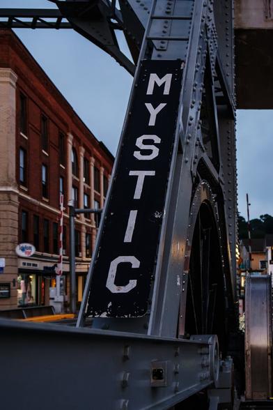 A color photograph of a portion of the upper structure of a drawbridge. A plate with the letters “MYSTIC” painted on it is mounted vertically on one of the upward sloping girders. A brick building is visible in the background, and the sky is a clear grayish-blue, just before daybreak.