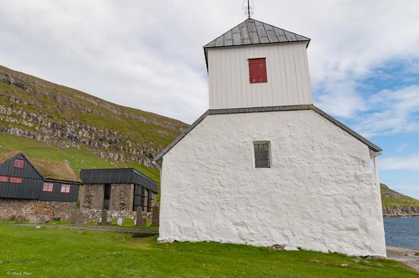 In the foreground is a small whitewashed stone church. From this angle there is a single small rectangular window set high in the wall of the church. Atop the building is a wooden tower which also contains a window but it has a red shutter closed across it.
Behind the church is the ruin of a medieval cathedral. All that largely remains are the stone walls and as part of a conservation project there is a protective shield on the top of the walls.
To the far left of the image is a wooden farmhouse with a turf roof and red painted window frames.
Behind all these buildings is a grass covered slope leading to a cliff face.